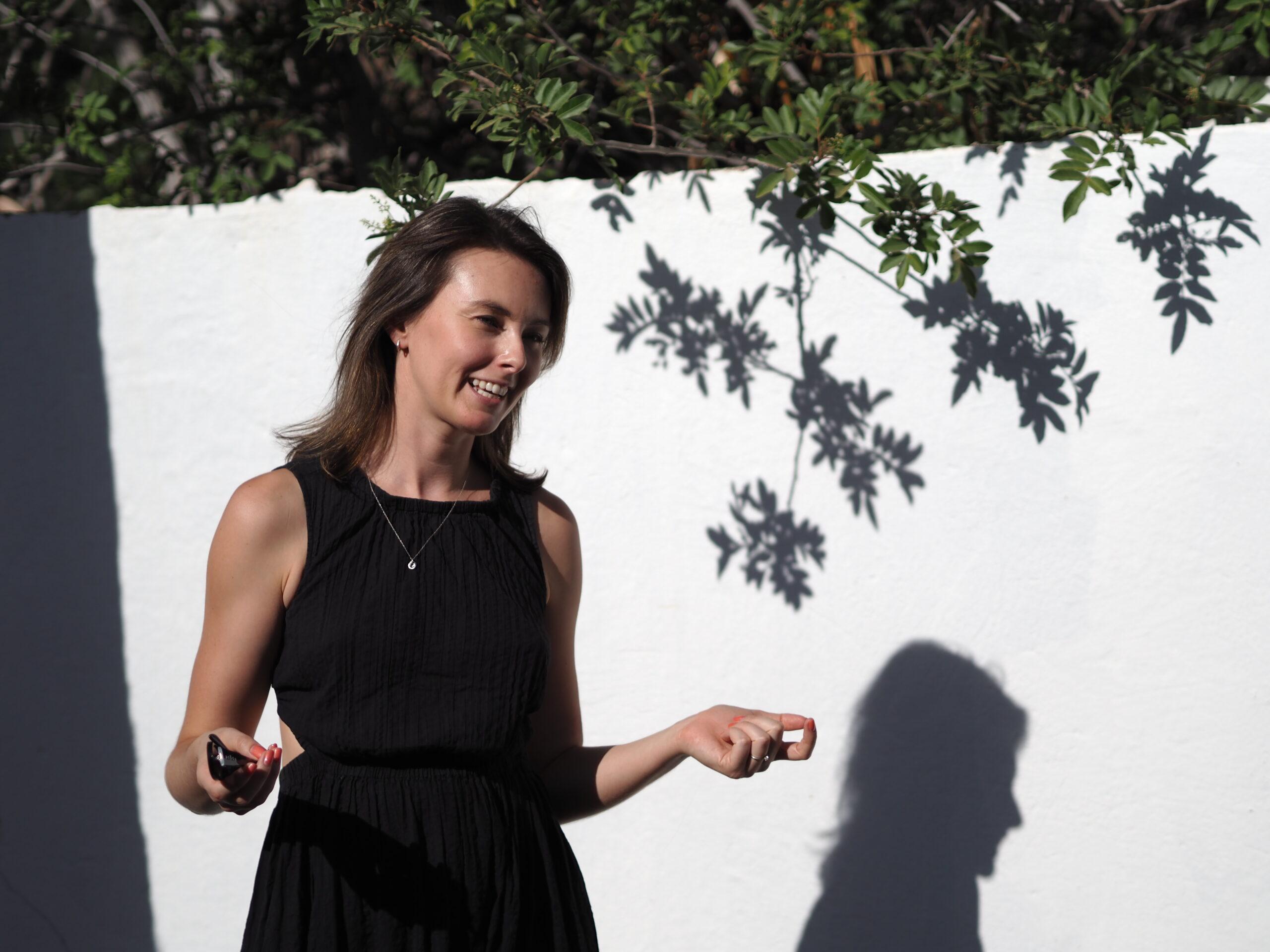 Becci smiling in front of a white wall with leaf shadows